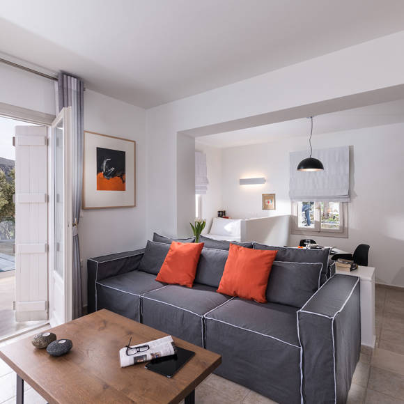 Junior villa living room with grey sofa, orange pillows, open kitchen and pool view terrace at Avdou Collection