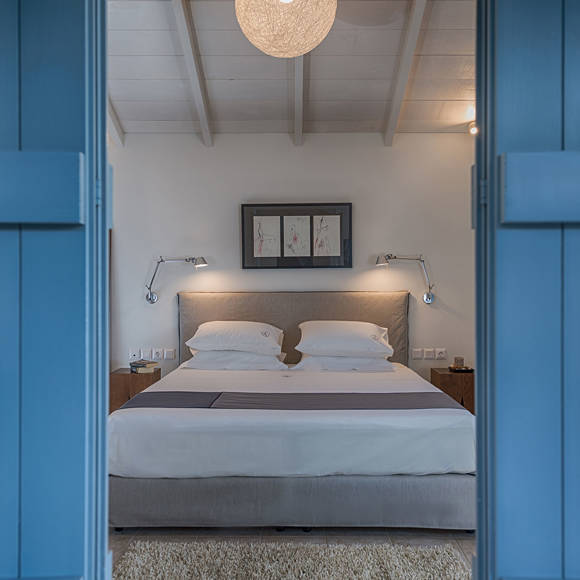 Junior villa bedroom with double bed, wall lamps, framed art and wooden nightstands viewd through blue doors at Avdou Collection Crete