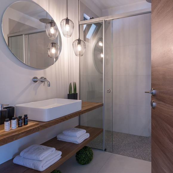 Grand villa bathroom with round mirror, glass shower, hanging lights and wooden vanity at Avdou Collection